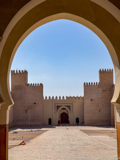 Fortified ceremonial gate with twin square towers and central doorway, seen through a large foreground arch across an open courtyard.
