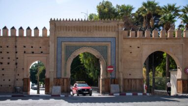 Decorated pointed arch in a crenellated Fes wall, with blue-green tilework, wooden doors, trees, and an adjacent smaller opening.