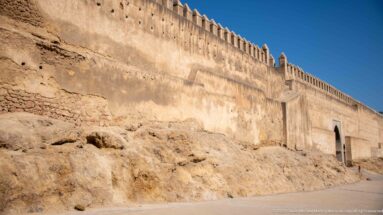Long crenellated earthen wall in Fes running above exposed rocky ground beneath a clear blue sky.