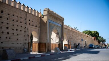 Long fortified wall in Fes with a decorated twin-arched gateway, wooden doors, crenellations, and a city street alongside.