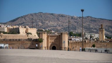 Wide paved square facing the crenellated walls and gateway of Fes, with towers and mountains in the background.