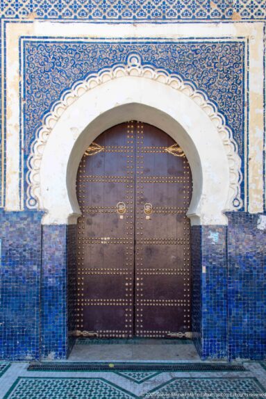 Dark studded wooden door inside a white horseshoe arch framed by blue tilework and geometric decoration.