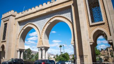 Large three-arched gateway over a road in Fes with carved geometric decoration, stone piers, blue window panels, cars, and palms.