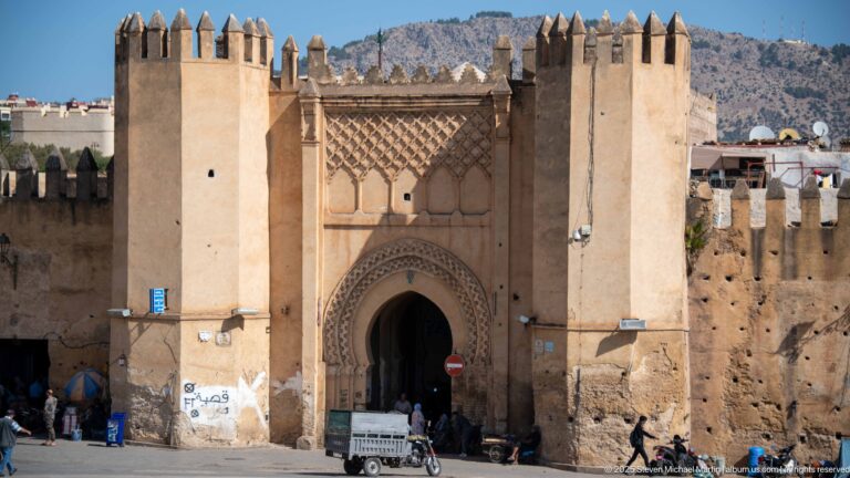 Historic Fes gate with pointed horseshoe entrance, flanking towers, crenellations, carved decoration, pedestrians, and a small vehicle.