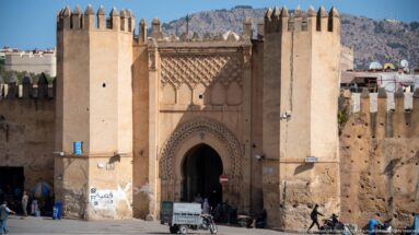 Historic Fes gate with pointed horseshoe entrance, flanking towers, crenellations, carved decoration, pedestrians, and a small vehicle.