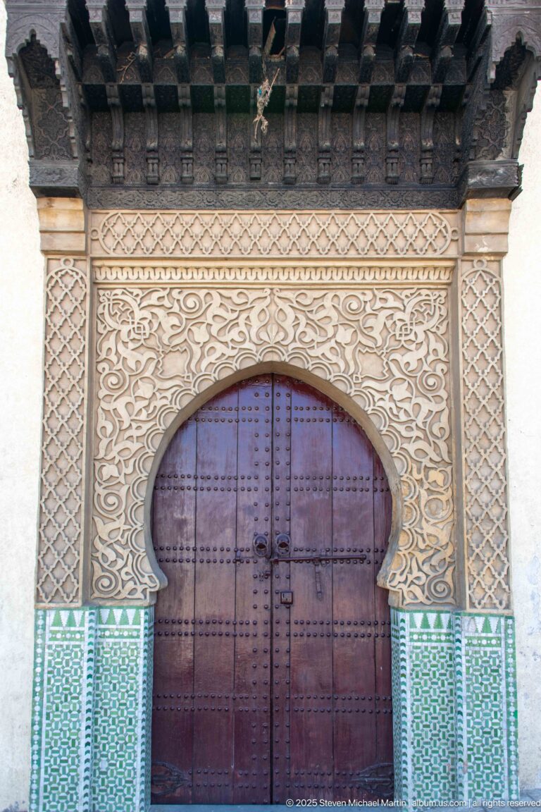 Large wooden door beneath a carved horseshoe arch, with geometric stucco borders, green zellij, and projecting timber beams.