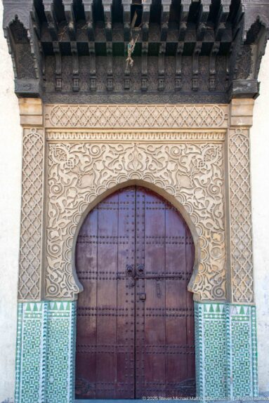 Large wooden door beneath a carved horseshoe arch, with geometric stucco borders, green zellij, and projecting timber beams.