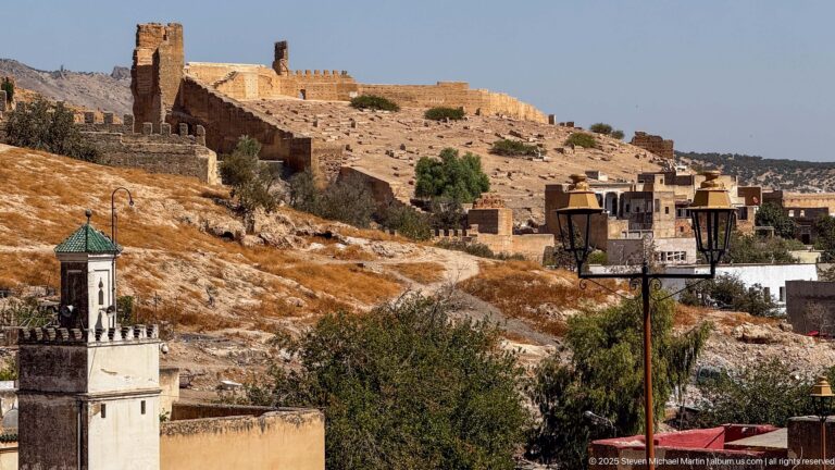 Historic earthen walls and square towers climb a dry hillside above Fes, with a small tower and modern buildings below.