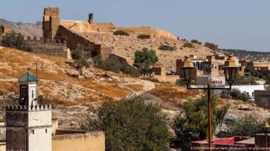 Historic earthen walls and square towers climb a dry hillside above Fes, with a small tower and modern buildings below.