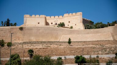 Borj Nord fortress above Fes, with pale defensive walls, corner towers, crenellations, and an open slope beneath a blue sky.