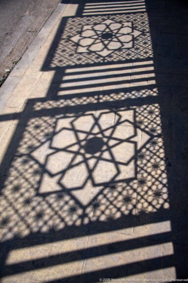 Sharp geometric lattice shadow with star patterns and horizontal bars falling across a stone pavement.