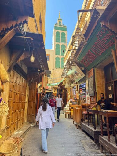 Narrow Fes medina street with shops and pedestrians leading toward a tall green-tiled minaret.