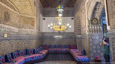 Corner of a Moroccan salon with low upholstered benches, zellij walls, carved decoration, hanging lamps, and a visitor at right.