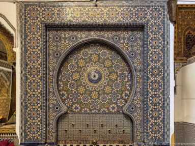 Large horseshoe-arched fountain niche covered in geometric zellij with a small basin and fittings at the base.