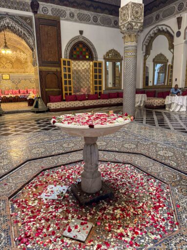 Interior courtyard with rose petals, central fountain, patterned rugs, carved columns, painted ceiling, and cushioned seating.