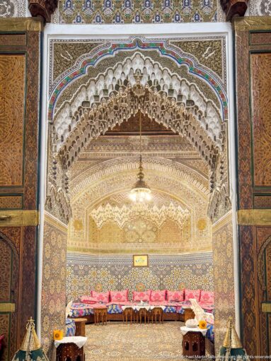 Muqarnas arch opening into a decorated salon with patterned tilework, painted wood, hanging lamp, and low upholstered seating.