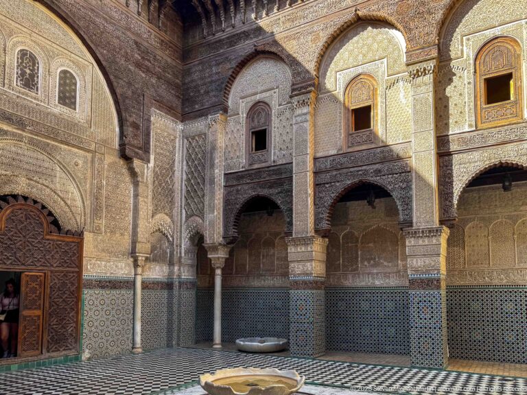 Arcaded Moroccan courtyard with zellij, carved plaster, columns, timber eaves, tiled floor, and a small central basin.