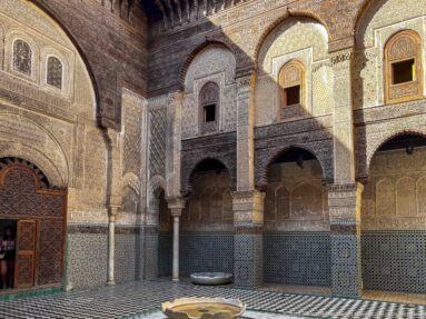 Arcaded Moroccan courtyard with zellij, carved plaster, columns, timber eaves, tiled floor, and a small central basin.
