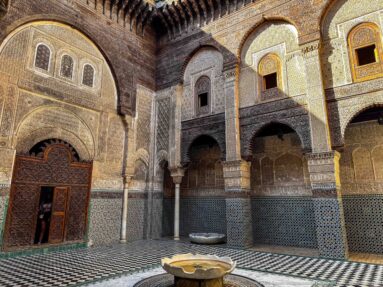 Wide view of an arcaded Fes courtyard with geometric tilework, carved plaster, timber eaves, doorway, and low fountain basin.