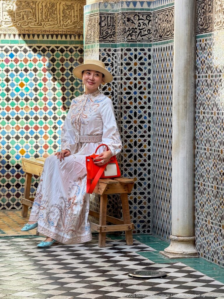 Seated woman in white clothing beside a wooden chair, framed by zellij, carved plaster, calligraphy, and a marble column.