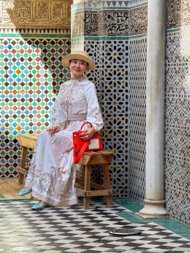 Seated woman in white clothing beside a wooden chair, framed by zellij, carved plaster, calligraphy, and a marble column.