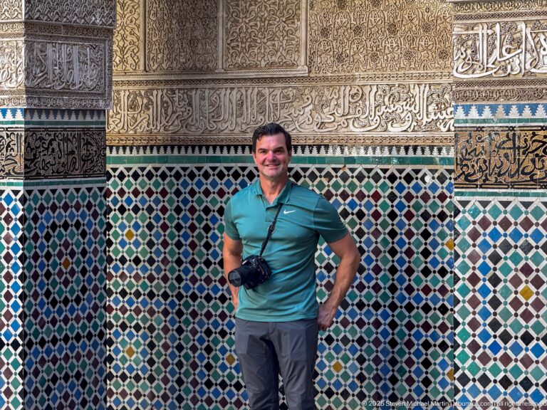 Traveler standing before multicolored zellij and carved plaster bands with Arabic calligraphy.