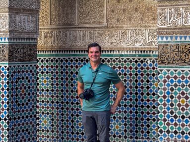 Traveler standing before multicolored zellij and carved plaster bands with Arabic calligraphy.