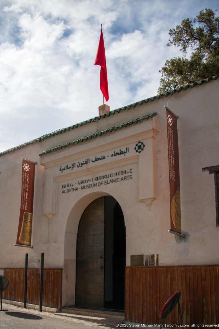 Pale museum façade with arched doorway, bilingual Al Batha signage, Moroccan flag, green-tiled roof edge, and two vertical banners.