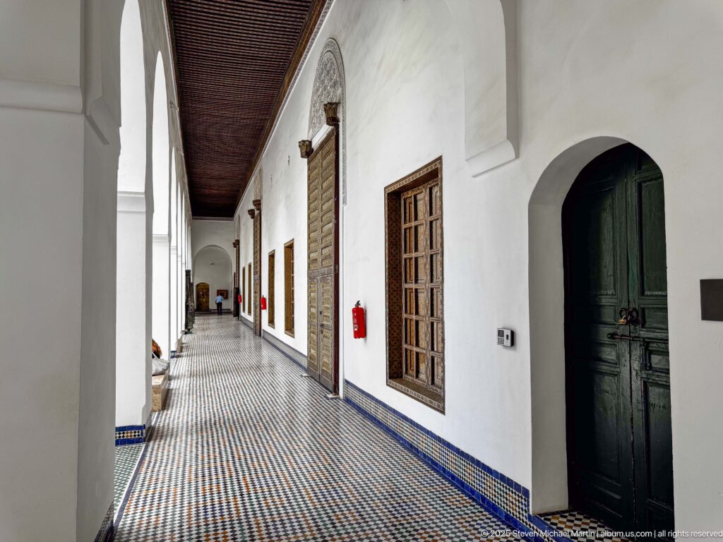 Long white museum arcade with repeated arches, carved wooden doors, grilled windows, blue tile edging, and patterned zellij floor.