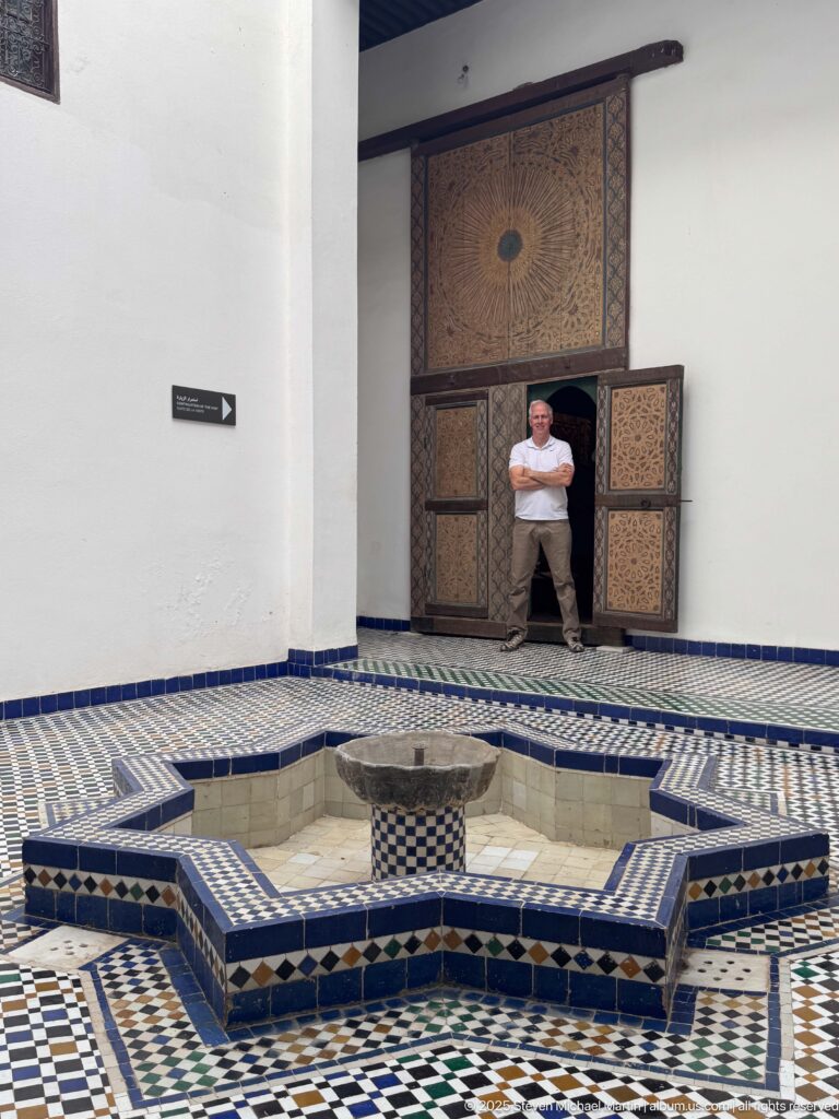 Nineteenth-century decorated wooden main door to Al Batha Palace’s throne hall, seen beyond the courtyard fountain with a visitor providing scale.