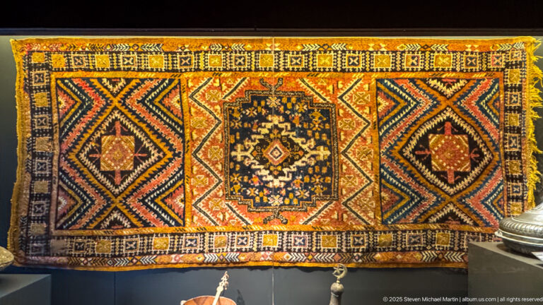 Rectangular woven textile with central dark medallion, red and black diamond fields, patterned borders, and yellow fringe.