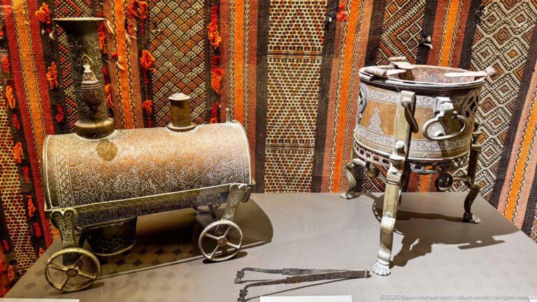 Ornamented cylindrical metal container on wheels beside a decorated round tripod vessel, displayed against patterned textiles.