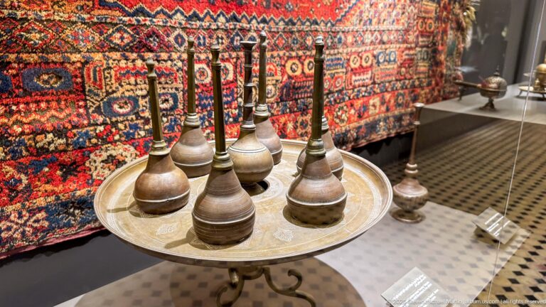Several bulbous metal vessels with long narrow necks arranged on a circular tray before a red patterned textile.