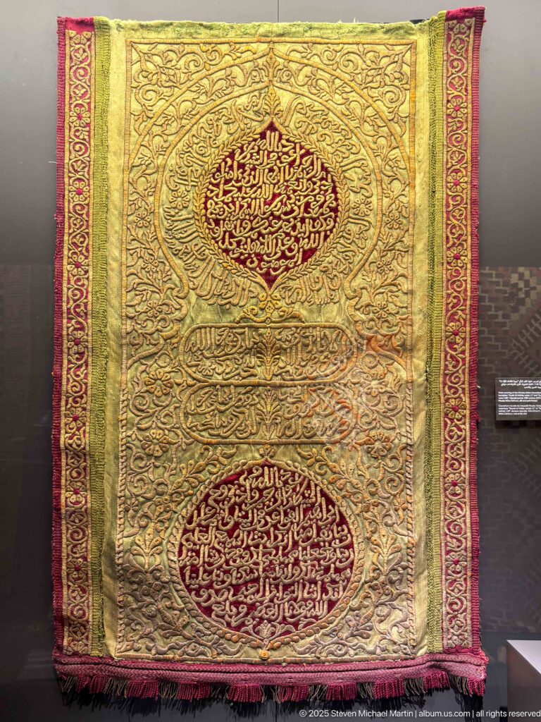 Gold-colored textile with two large red Arabic calligraphic medallions, scrolling ornament, and narrow red side borders.