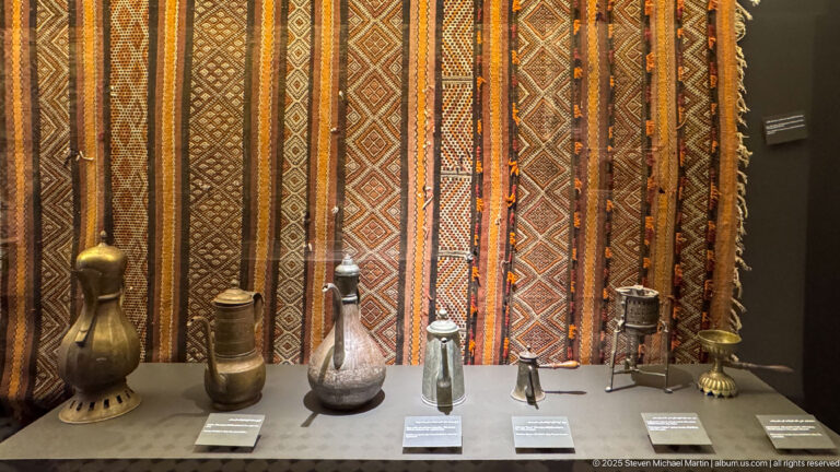 Row of engraved metal ewers, bottles, and containers displayed against vertically hung orange, brown, and black woven textiles.