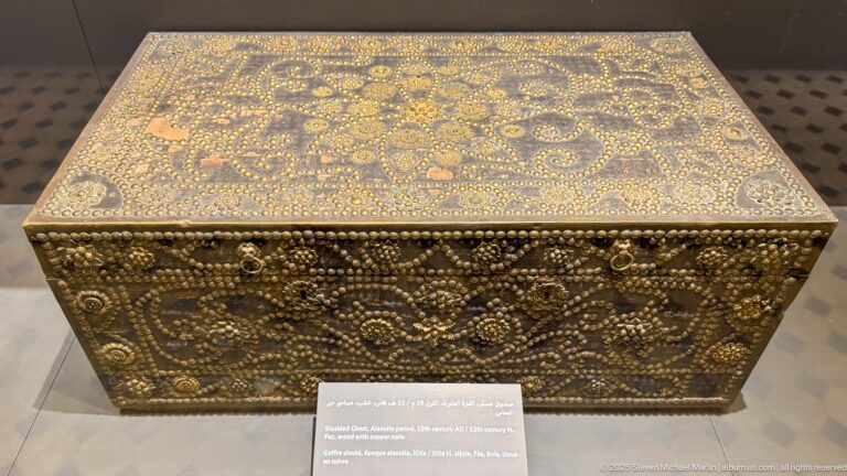 Rectangular dark wooden chest densely decorated with brass-colored studs forming scrolls, rosettes, borders, and corner motifs.