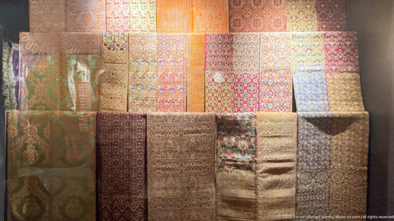 Large museum display of folded and hanging textiles in red, gold, green, blue, cream, and brown with varied repeating patterns.
