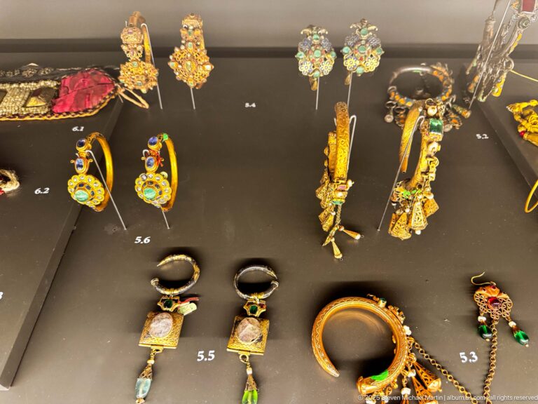 Assorted gold-colored earrings, rings, bracelets, and small ornaments set with red, green, blue, and turquoise stones on a dark display.