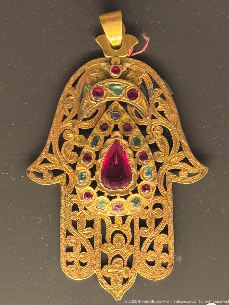 Gold-colored openwork hamsa hand pendant with a large red teardrop stone and smaller red, green, and blue settings.