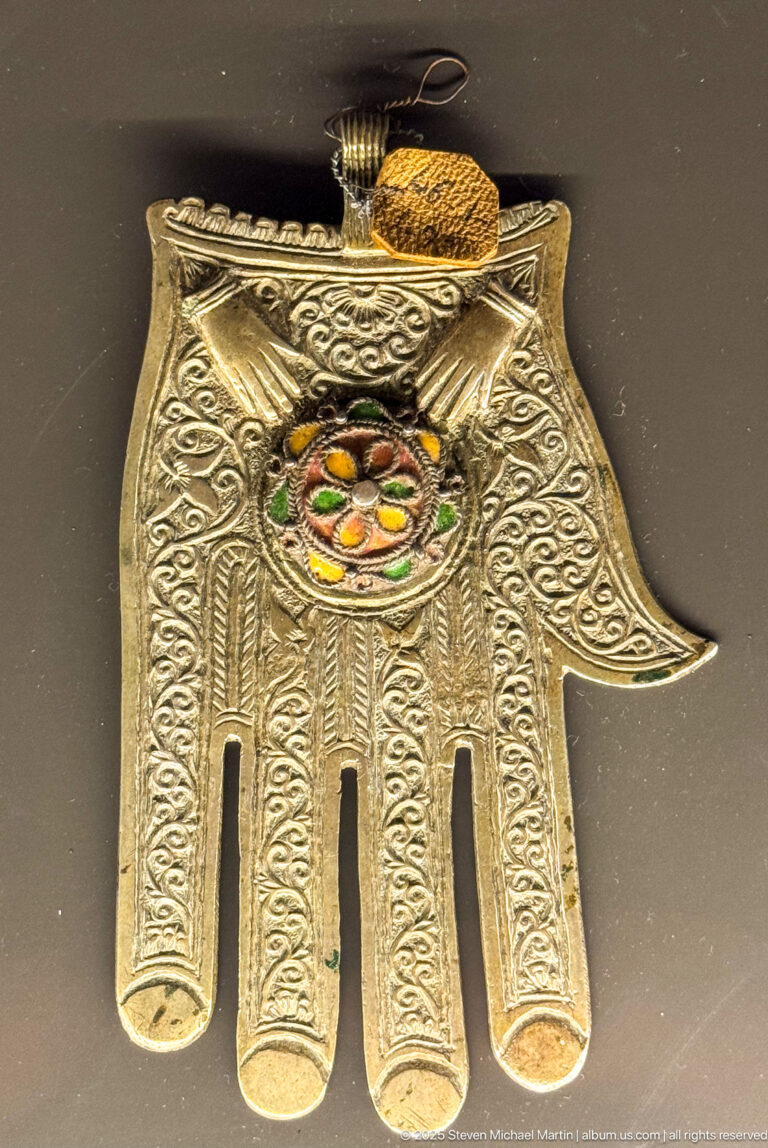 Silver-colored hand-shaped hamsa amulet with engraved scrollwork, four finger openings, suspension loop, and multicolored central setting.