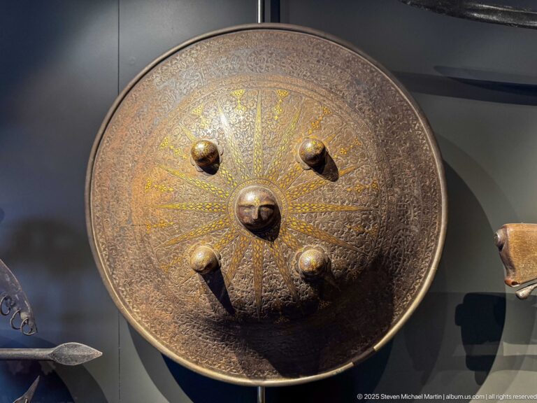 Circular engraved metal shield with a large central boss, four smaller bosses, radiating ornament, and traces of gold-colored decoration.