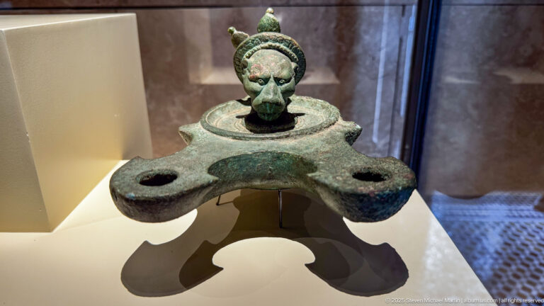 Patinated bronze oil lamp with three projecting nozzles, central reservoir, hinged lid, and small figural finial.