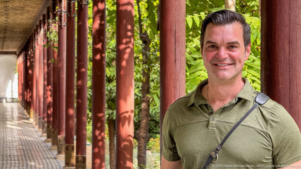 Visitor in a green shirt standing beside red wooden columns along a shaded garden arcade with dense foliage behind.