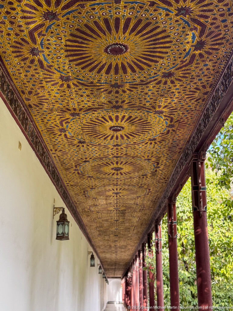 Long covered gallery with richly painted wooden ceiling of radiating stars, red columns, white wall, lanterns, and garden foliage.