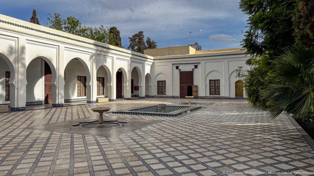 White arcaded courtyard with extensive patterned tile paving, a pedestal fountain, rectangular basin, dark wooden doors, and palms.