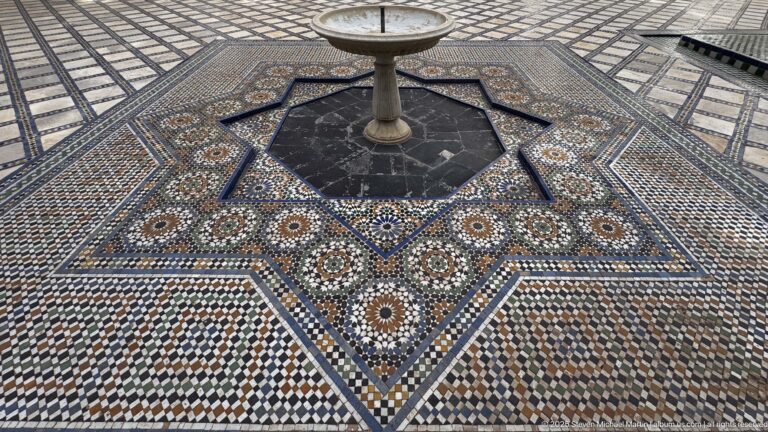 Pedestal fountain in a dark octagonal pool surrounded by star-shaped zellij with circular rosettes and multicolored geometric borders.