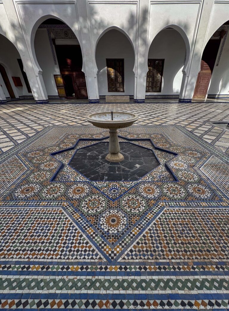 Low view across multicolored star-pattern zellij toward a pedestal fountain, dark octagonal pool, and white courtyard arcade.