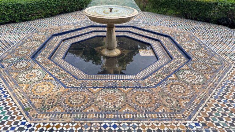 Octagonal tiled fountain with central pedestal, dark reflective water, concentric zellij borders, rosettes, and clipped hedges behind.