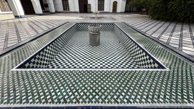 Large rectangular sunken basin lined with green, white, and black geometric tile, with a cylindrical stone element near the center.