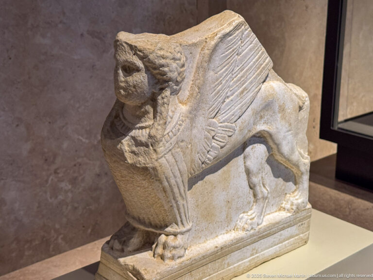 Carved stone winged quadruped with raised head, feathered wings, four legs, and rectangular base in a museum case.